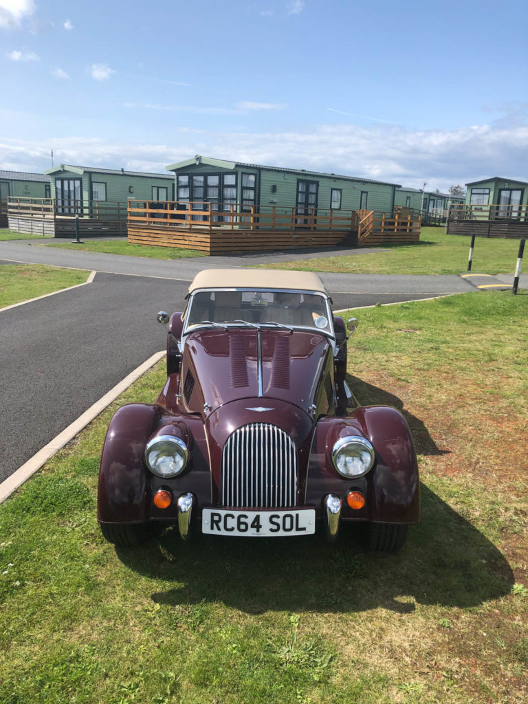 Morgan Plus 4 parked on grass verge in front of static caravans