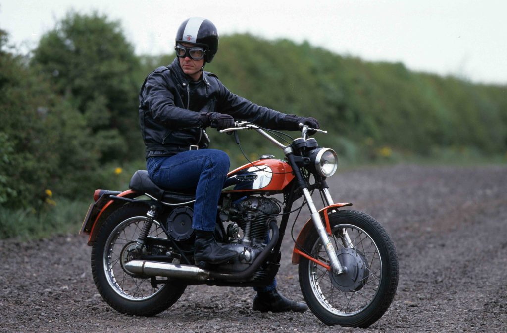 Rider sitting on red and silver classic motorbike parked on gravel road