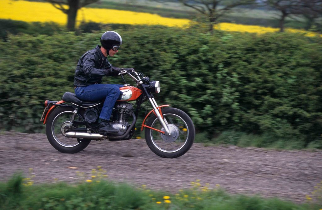 Man riding red and silver classic motorbike on gravel road through countryside