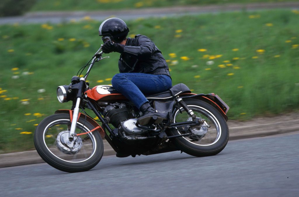 Man riding red and silver classic Ducati 350 Scrambler motorbike on tarmac road through countryside