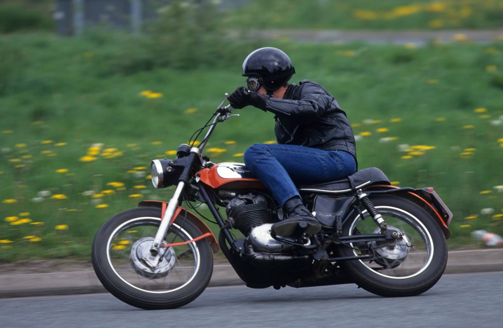 Man riding red and silver classic Ducati 350 Scrambler motorbike on tarmac road through countryside