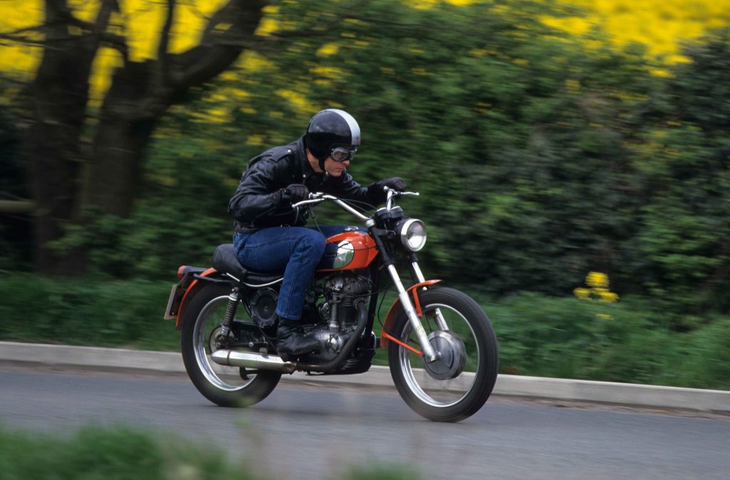 Man riding red and silver classic motorbike on tarmac road through countryside