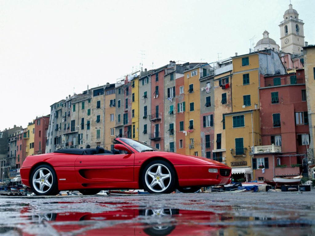 Ferrari F355 Spider parked on wet tarmac with yellow and red high-rise buildings behind 