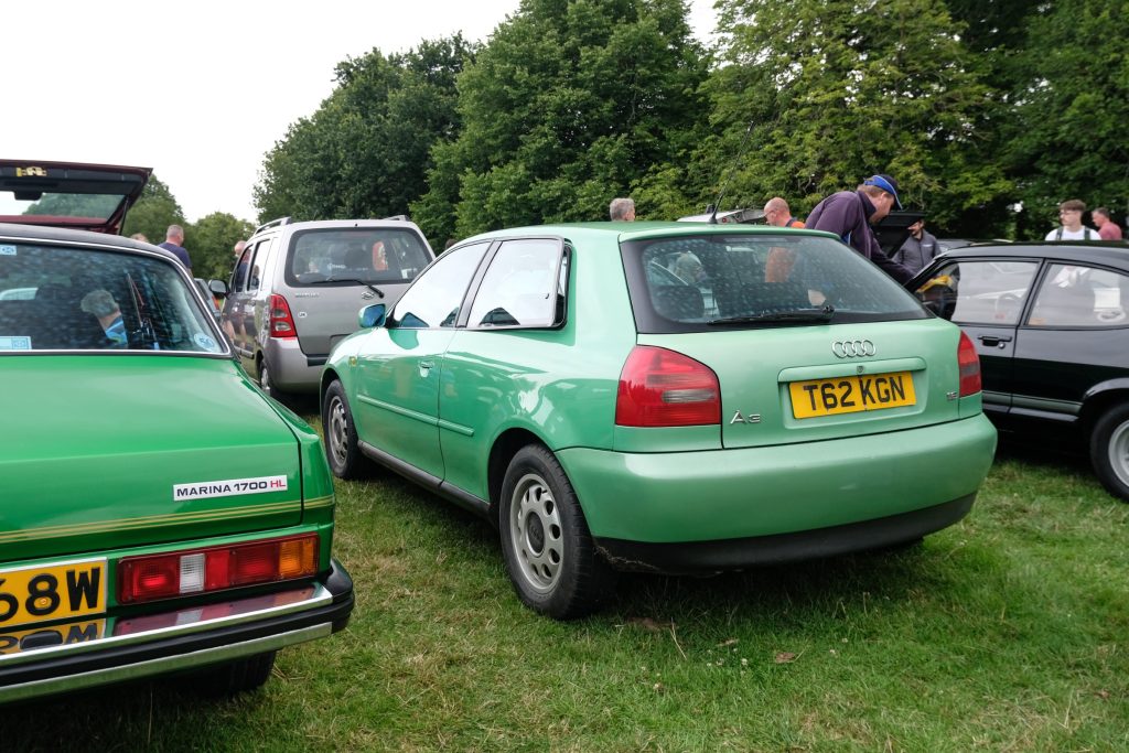 Several cars parked in a row in grass field with trees behind