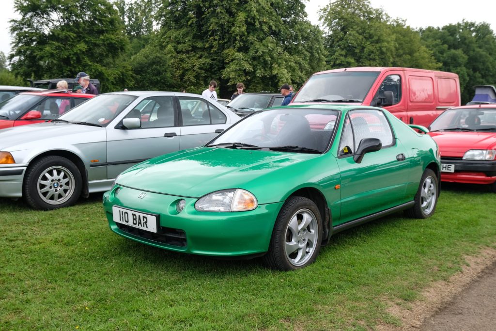Several cars parked in a row in grass field with trees behind