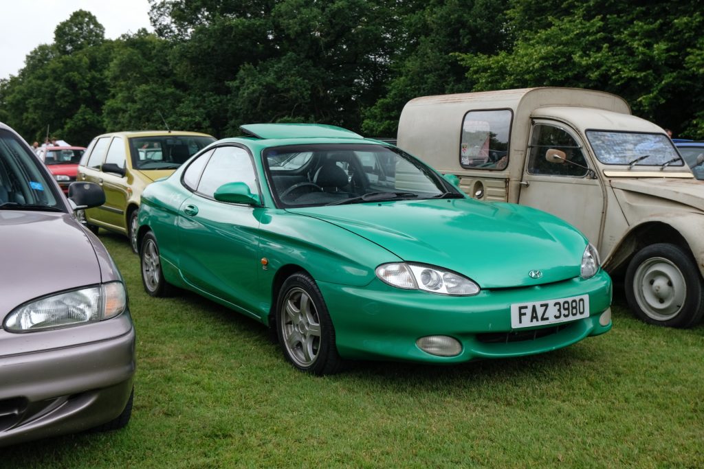 Several cars parked in a row in grass field with trees behind