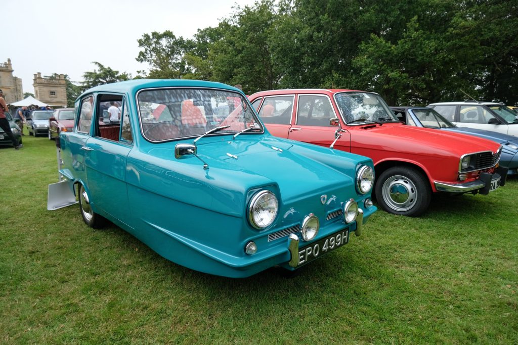 Several cars parked in a row in grass field with trees behind
