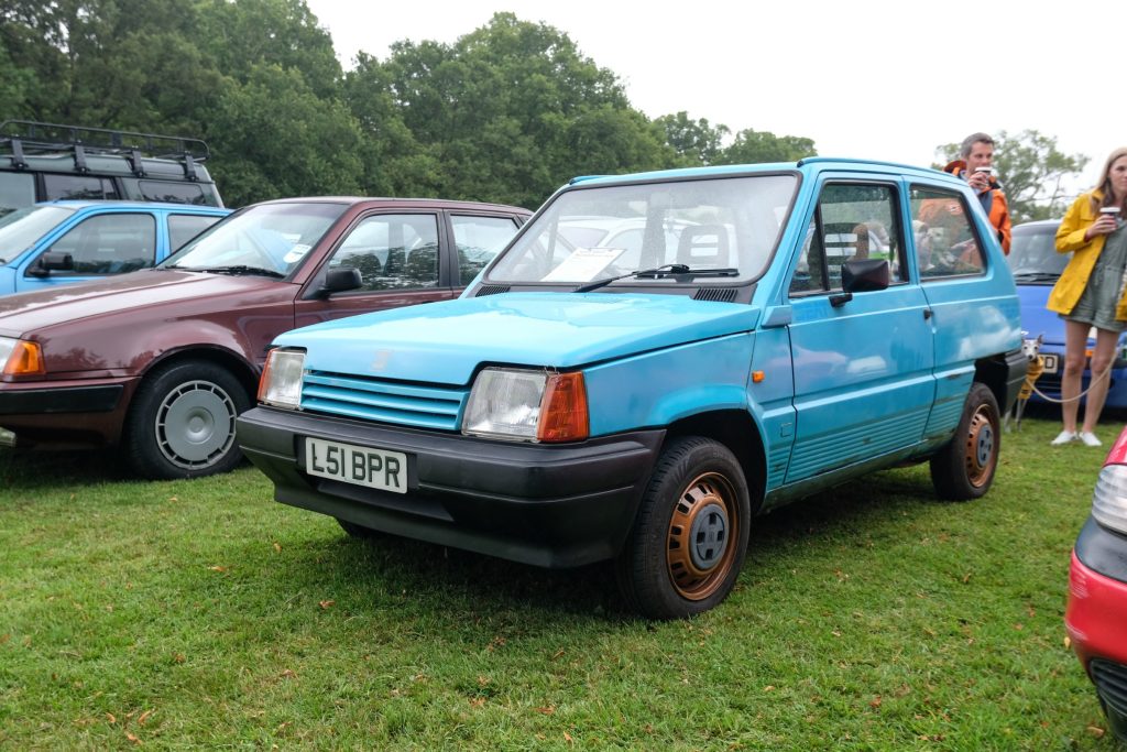 Several cars parked in a row in grass field with trees behind