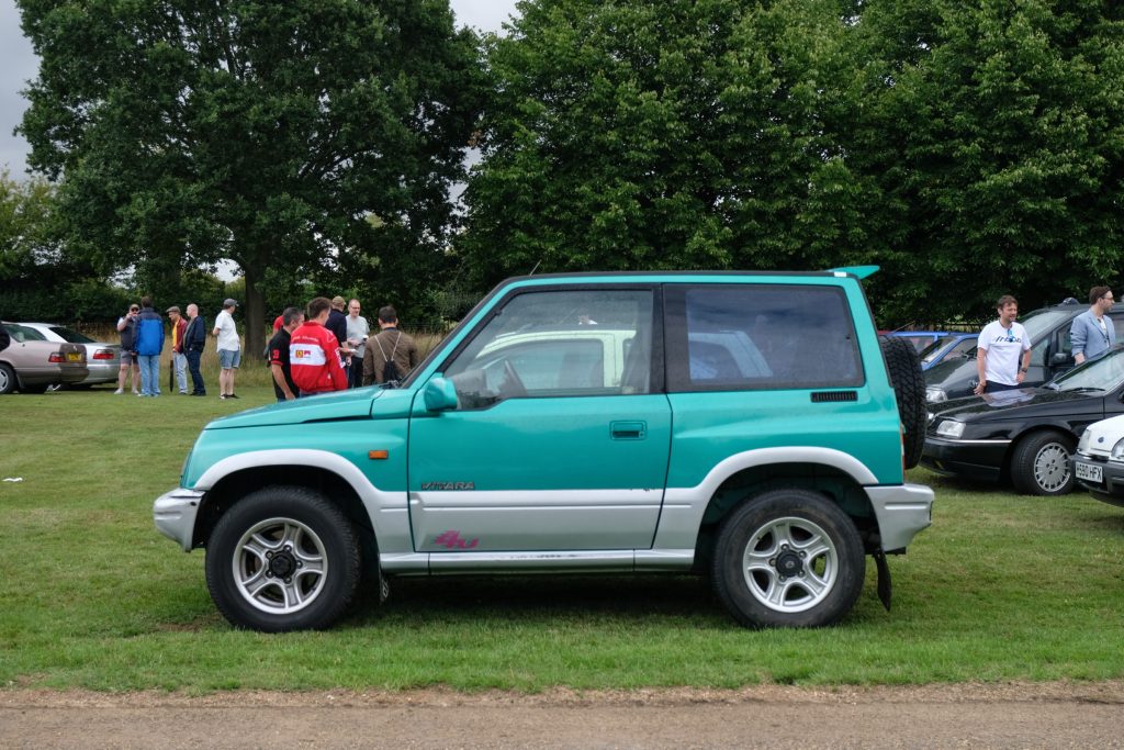 Several cars parked in a row in grass field with trees behind