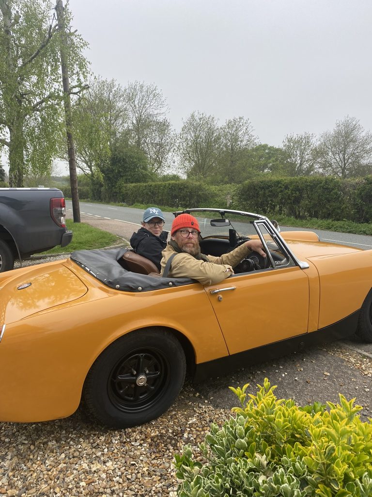 Yellow open-top classic car parked on a driveway with a passenger and a driver inside
