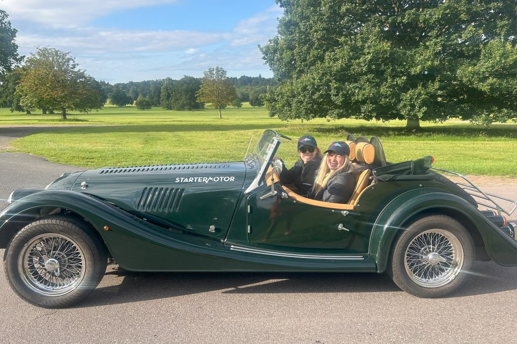 Two women sat inside open open-top classic car parked in front of fields and trees