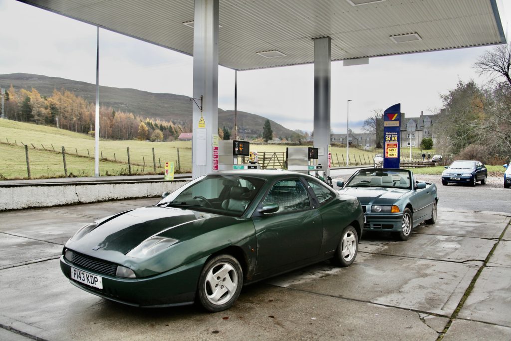 Two classic cars parked in a fuel station forecourt