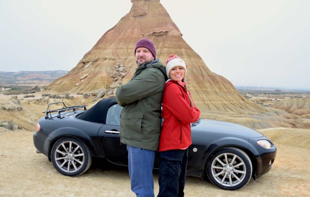 Two people posing back to back in front of car and mountain