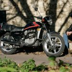 Man sat next to red and black motorbike parked up in front of a stone wall