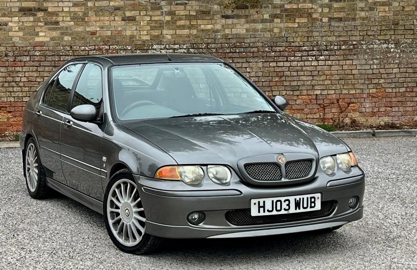 Grey MG car parked on gravel surface with red brick wall behind