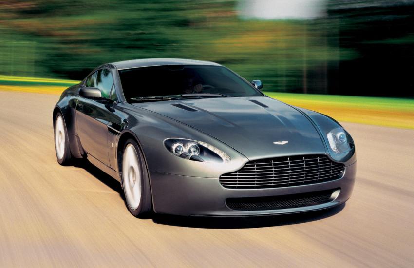 Front view of a greyAston Martin Vantage driving along a road with blurred trees behind