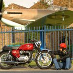 Man sat next to parked BSA Spitfire MkIV motorbike in front of a Spitfire plane