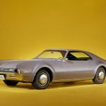 Side profile of grey Oldmobile Toronado on yellow background