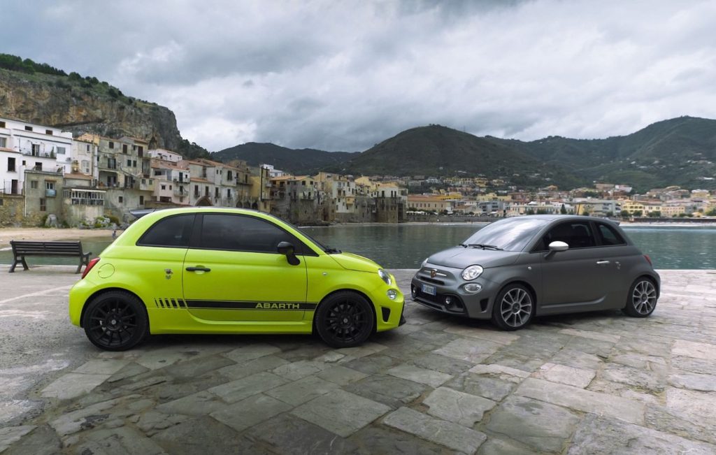 Two Abarth 595 Competizione hatchback cars in different colors, set against a coastal Italian town backdrop