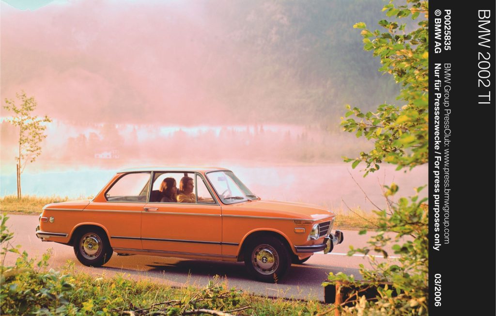 Man and woman sat inside an orange classic car parked at the side of a misty lake