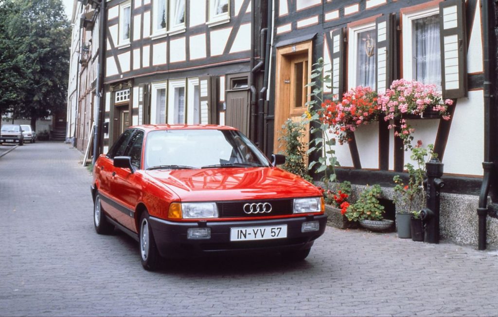 Red Audi 80 parked on a road next to a black and white tudor-style building