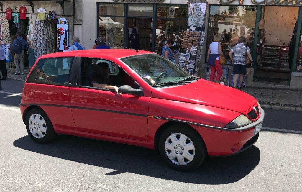 A red Lancia Ypsilon (Type 840) parked or moving slowly near storefronts.