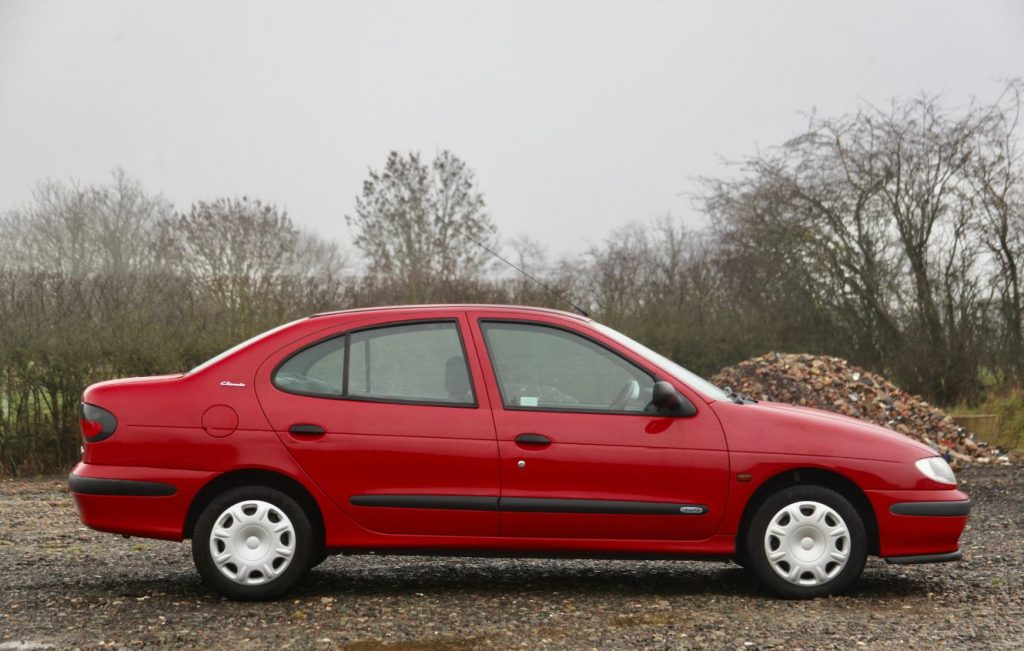 Side view of a red Renault Megane parked on gravel in front of trees on a cloudy day