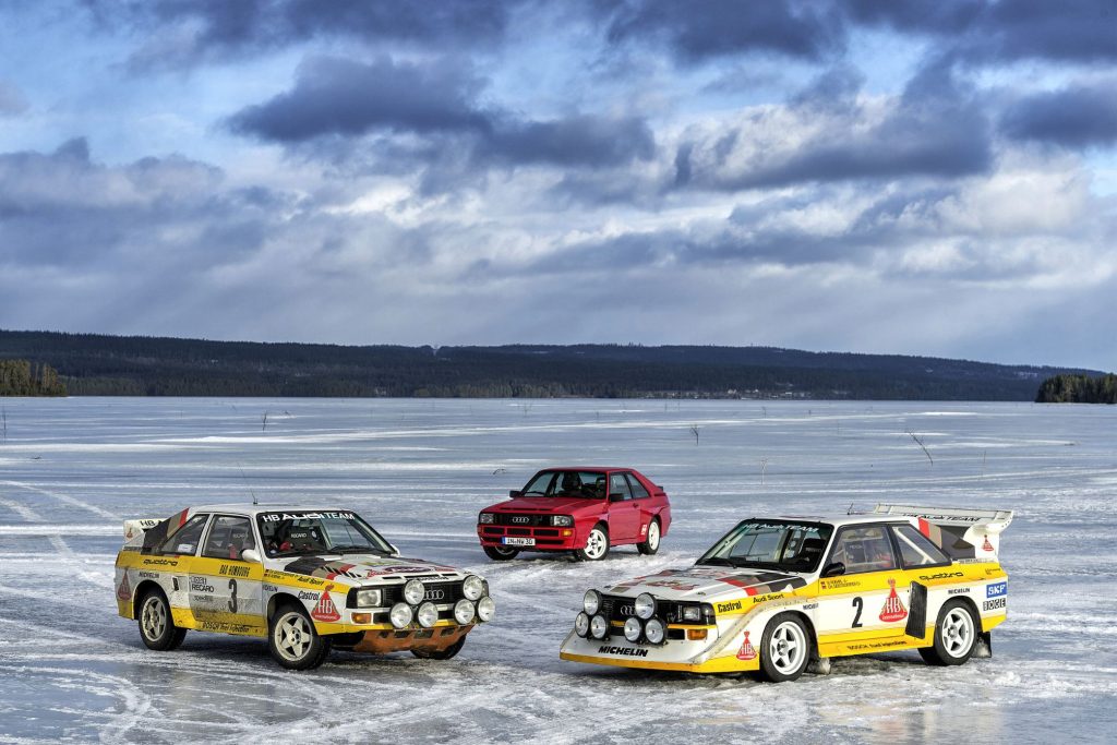 Two Audi Sport Quattro Group B rally cars in HB livery parked on a frozen lake alongside a red road-going Audi Quattro