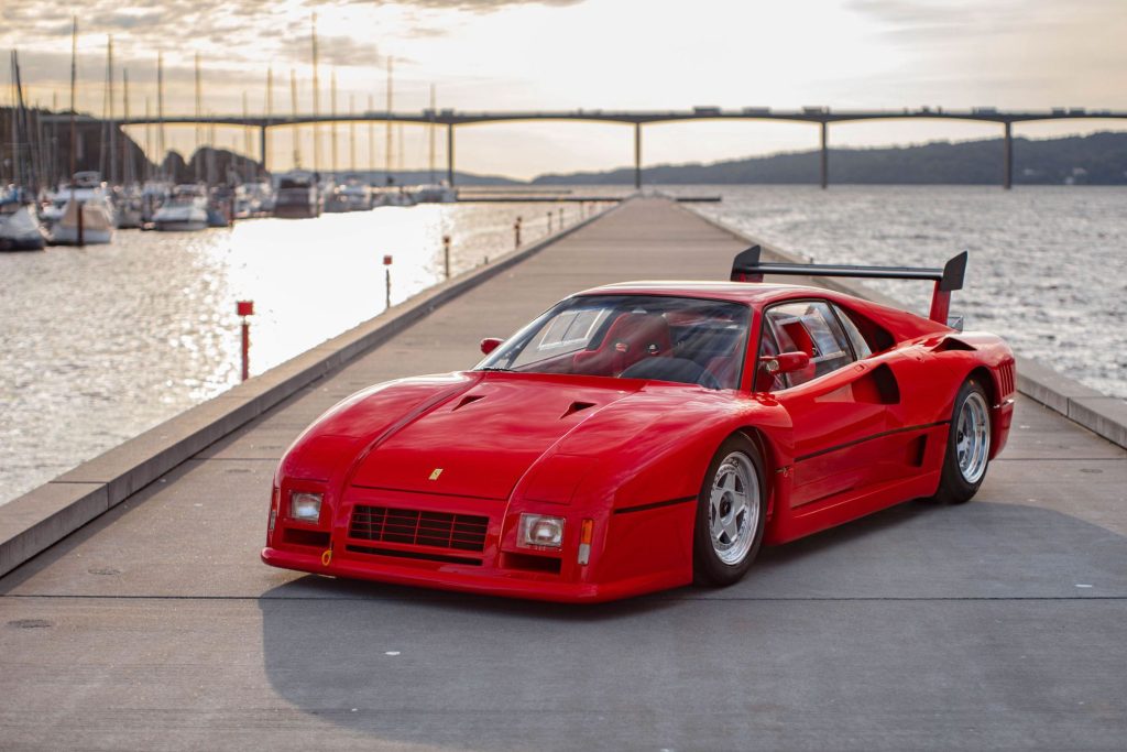 Red Ferrari 288 GTO Evo Group B car parked on a harbourside road at dusk with boats and a bridge in the background