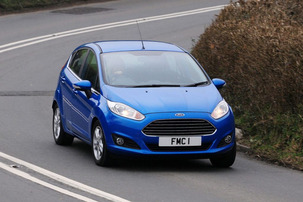 Blue Mk7 Ford Fiesta five-door hatchback being driven on a country road past a hedgerow