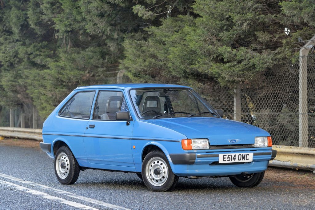 Blue Mk2 Ford Fiesta three-door hatchback parked on a wet road in front of trees and a wire fence