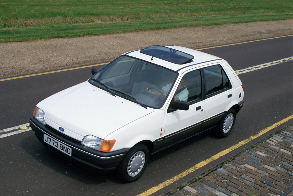 White Mk3 Ford Fiesta five-door hatchback with a sunroof, photographed from above on a road with yellow lines and a cobbled edge