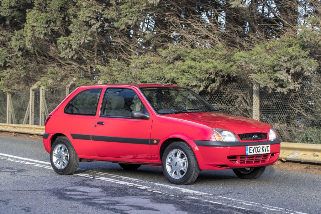 Red Mk5 Ford Fiesta three-door hatchback parked on a wet road in front of trees and a wire fence