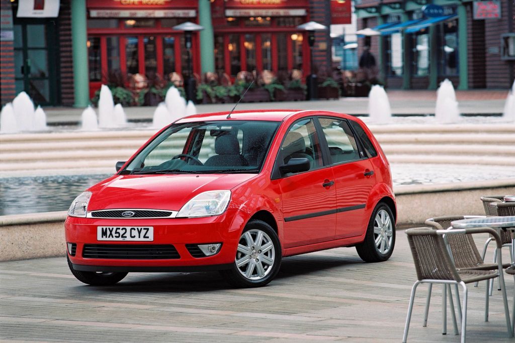 Red Mk6 Ford Fiesta five-door hatchback parked in a town square with water fountains and café seating in the background