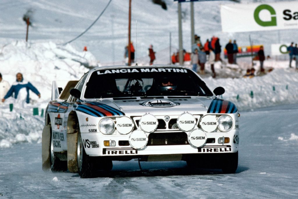 Lancia 037 Rallye in Martini Racing livery on an icy stage with spectators and snow banks in the background