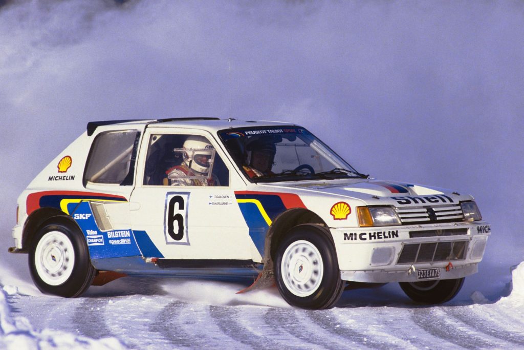 Peugeot 205 T16 Group B rally car in blue and white livery sliding on a snowy rally stage with the driver and co-driver visible