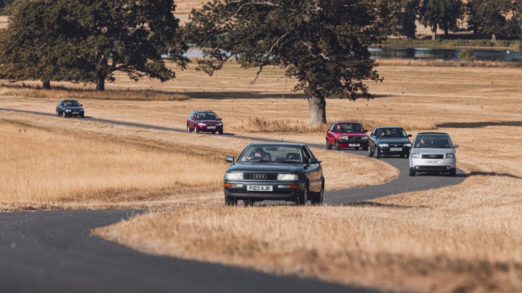 Classic cars driving long a winding road through a field with trees and a lake in the background arriving first at FOTU