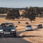 Classic cars driving long a winding road through a field with trees and a lake in the background