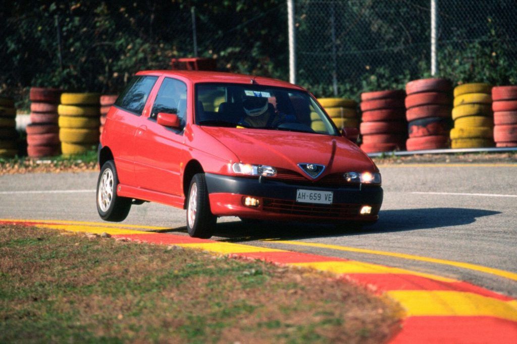 Red Alfa Romeo 145 Quadrifoglio three-door hatchback being driven on a race circuit with painted tyre barriers visible in the background