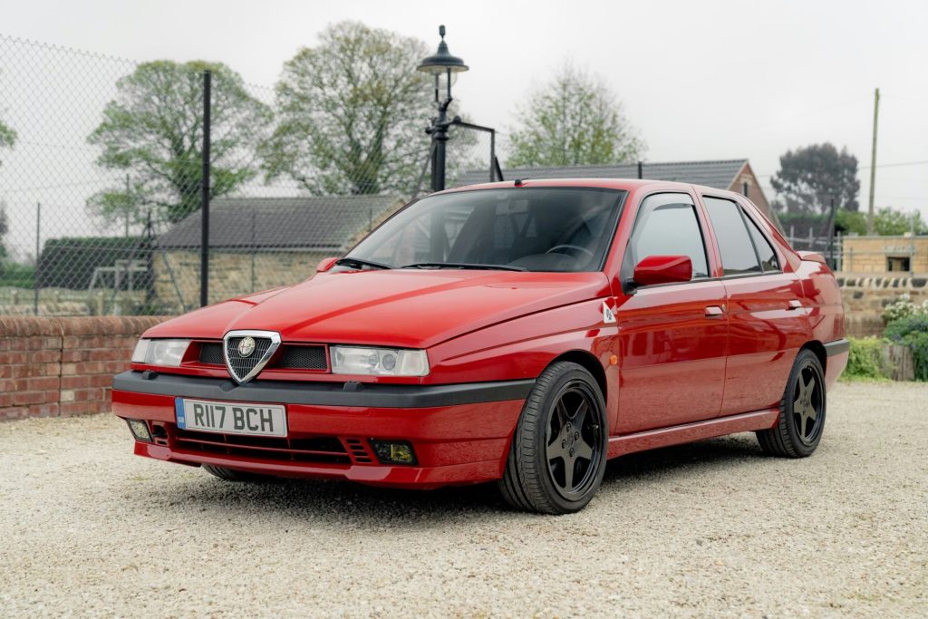 Red Alfa Romeo 155 saloon with black alloy wheels parked on gravel in front of a stone wall and wire fence