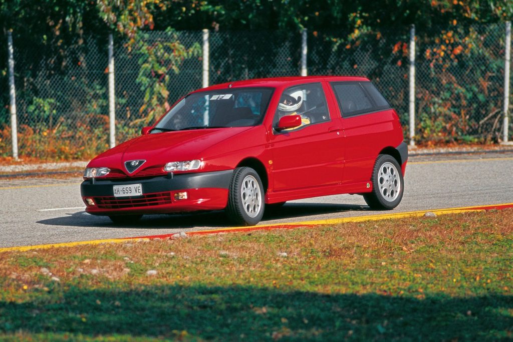 Red Alfa Romeo 145 Quadrifoglio three-door hatchback being driven on a race circuit with a helmeted driver visible