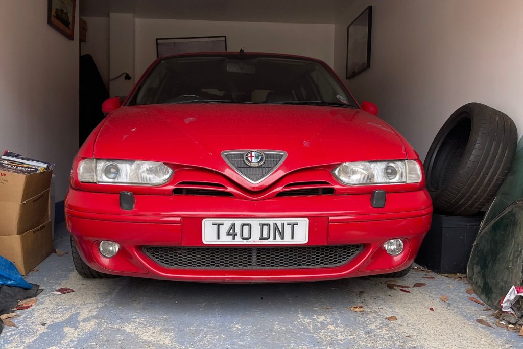 Red Alfa Romeo 145 Cloverleaf parked head-on in a domestic garage with spare tyres and cardboard boxes alongside
