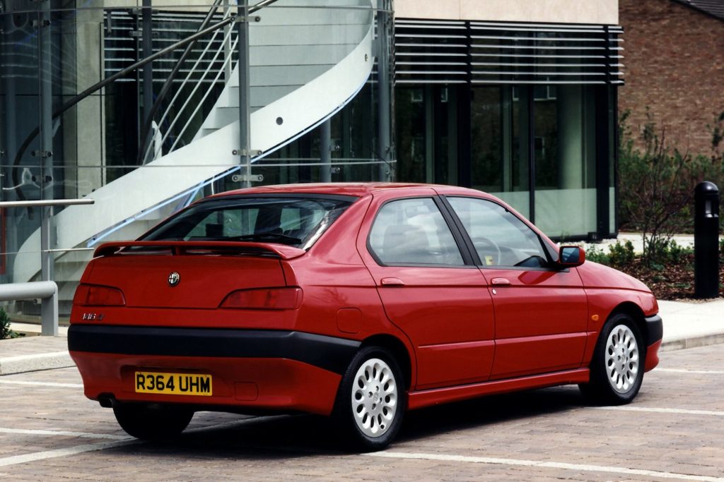 Rear three-quarter view of a red Alfa Romeo 146 Ti saloon parked on cobblestones outside a modern glass building