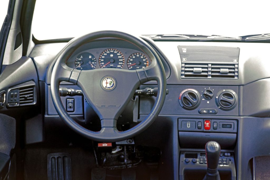 Alfa Romeo 145 dashboard and steering wheel interior shot showing instrument cluster, climate controls and red-stitched gear knob