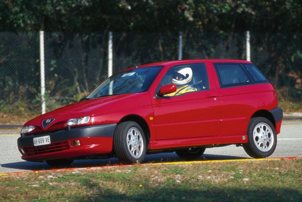 Red Alfa Romeo 145 Quadrifoglio three-door hatchback in side profile on a race circuit with a helmeted driver