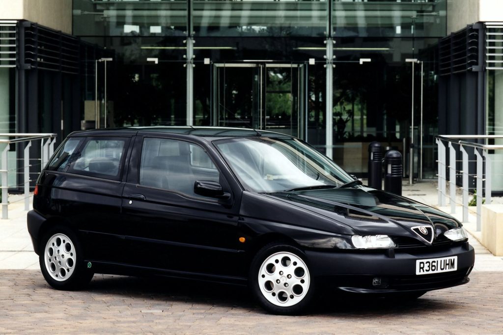 Black Alfa Romeo 145 Cloverleaf five-door hatchback parked on cobblestones outside a modern glass building
