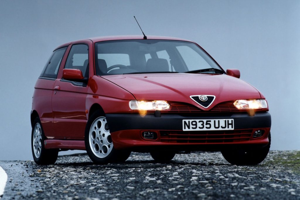 Red Alfa Romeo 145 Cloverleaf three-door hatchback photographed head-on with headlights on, parked on gravel against a pale sky