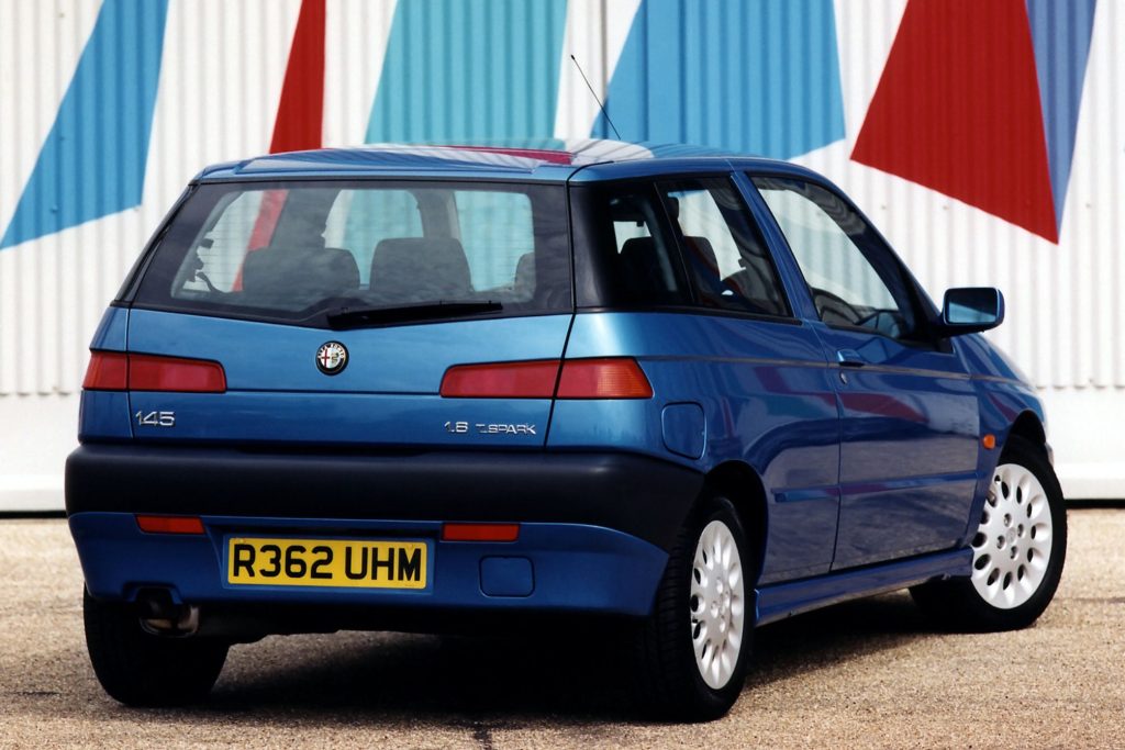 Rear three-quarter view of a blue Alfa Romeo 145 1.8 Twin Spark three-door hatchback parked in front of a colourful striped wall