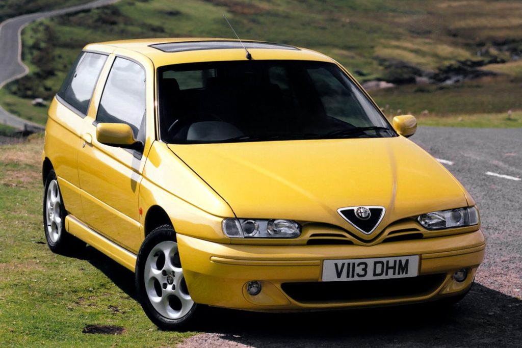 Yellow Alfa Romeo 145 three-door hatchback parked on a hillside road with green hills and a winding road in the background