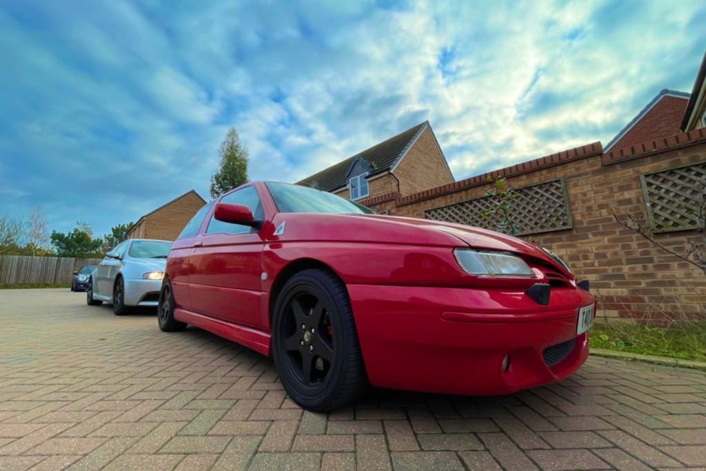 Low angle front three-quarter view of a red Alfa Romeo 145 Cloverleaf with black alloys parked on a brick driveway with houses and a dramatic cloudy sky behind